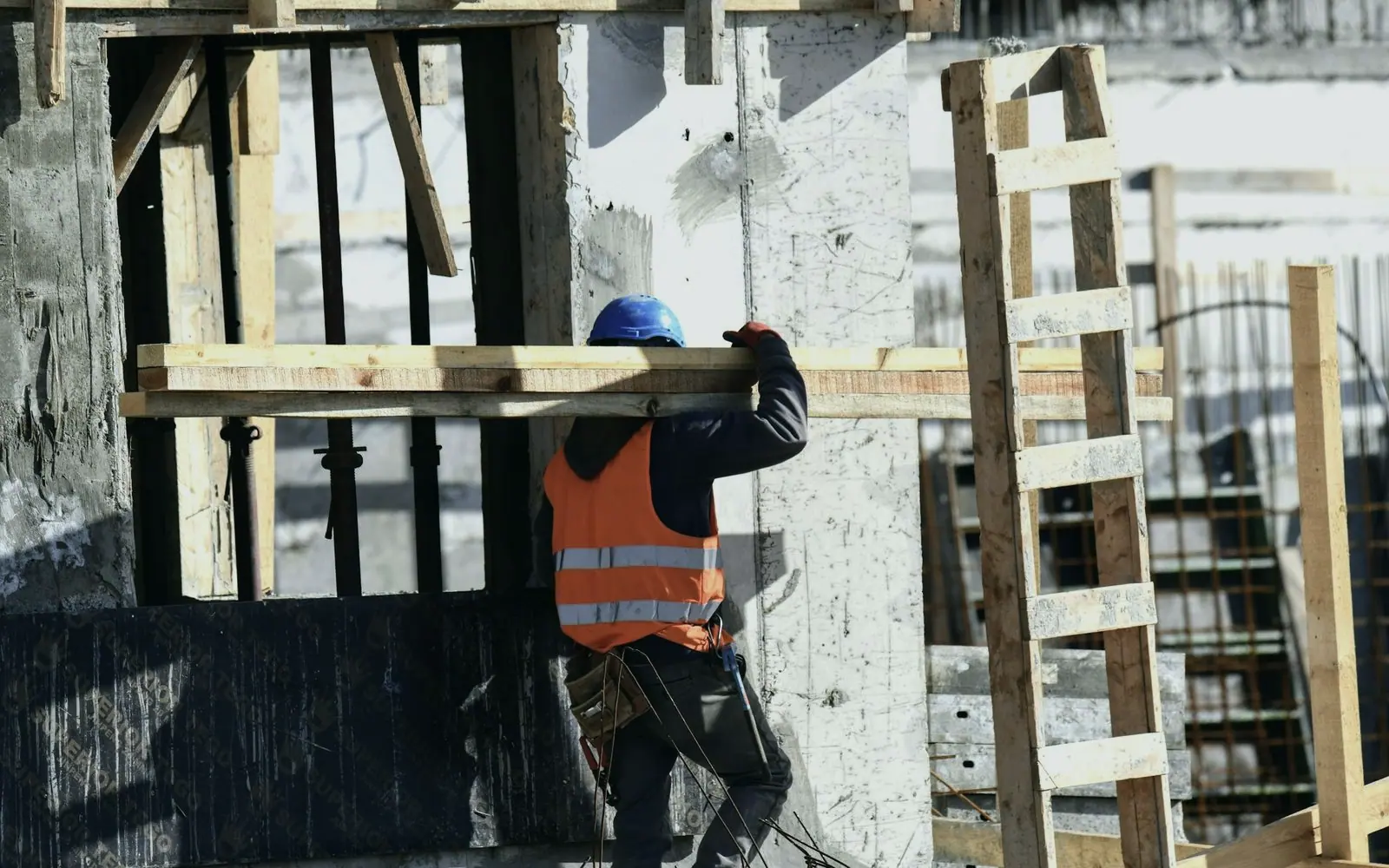 A construction worker in a hard hat and high-visibility vest carrying timber on site