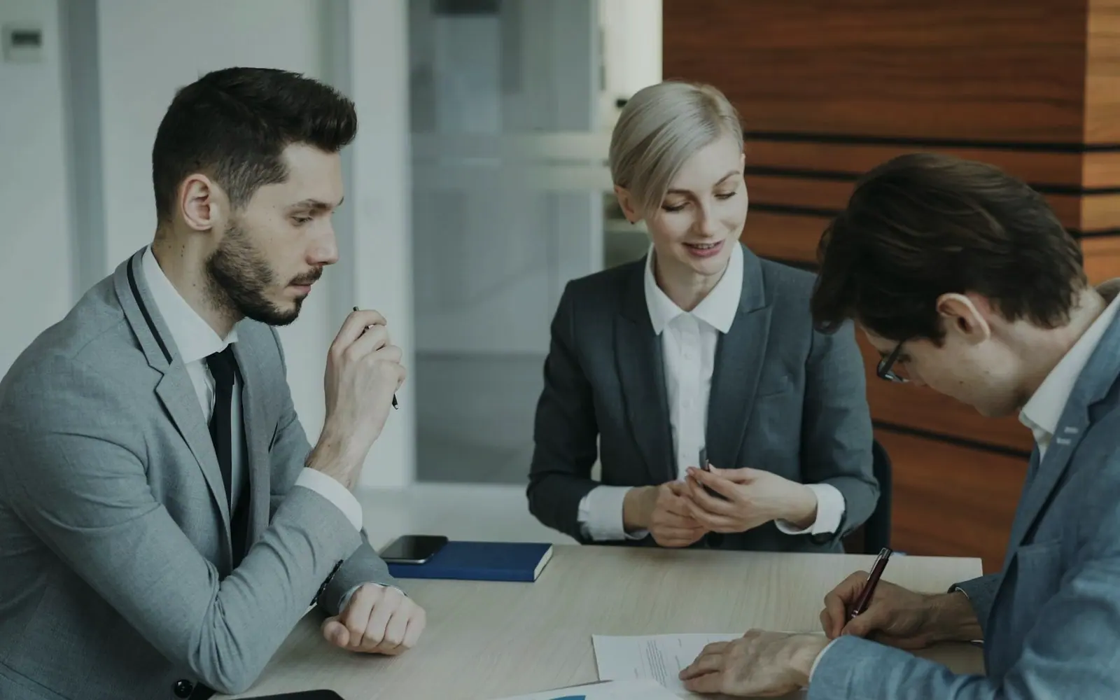 Three colleagues in suits reviewing and signing a contract at a table