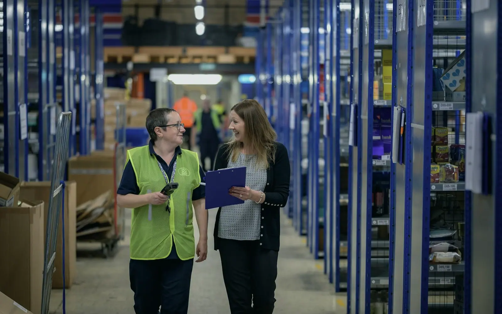 Two colleagues in high-visibility vests walking a warehouse aisle